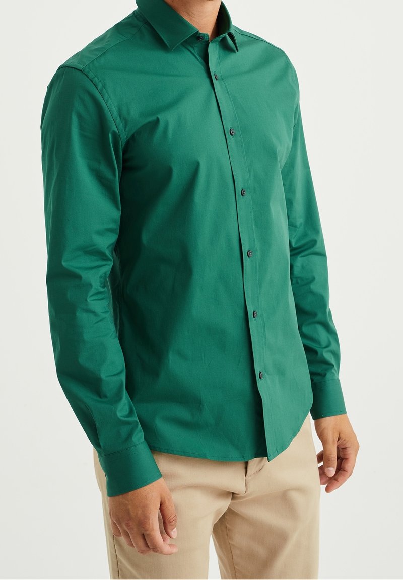 Grön skjorta med knappar i en mjuk tyg, med långa ärmar, spetsig krage och svarta knappar fram, tillsammans med beige byxor.