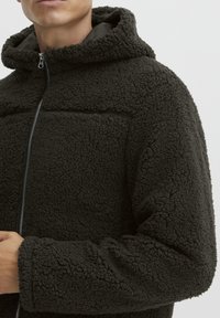 Ciemnozielona bluza polarowa z teksturowaną, puszystą powierzchnią; ma kaptur, pełny zamek błyskawiczny i minimalistyczny design bez widocznego logo.