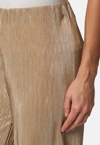 Pantalons plissés en métal doré avec une surface texturée et une taille ajustée, mettant en avant des rayures verticales et un éclat lisse.
