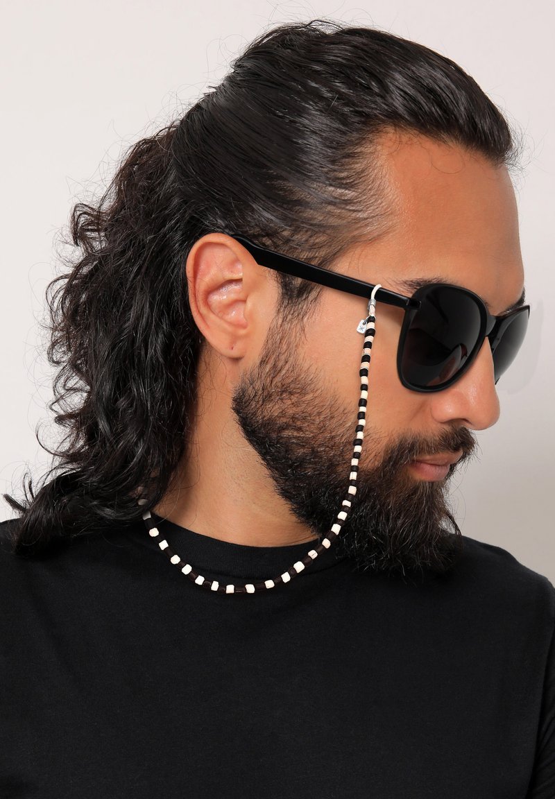 Mann mit langen dunklen Haaren und Bart, der schwarze Sonnenbrille mit einer schwarz-weißen Perlenbrille trägt und ein schwarzes Hemd anhat.