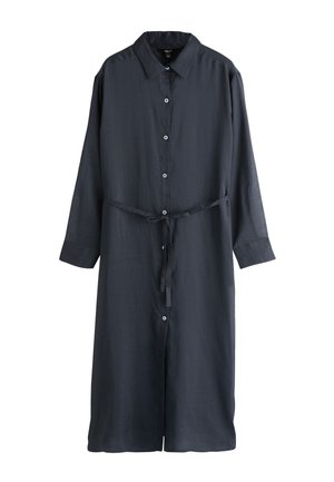 Vrouw in een lange zwarte geknoopte jurk en sandalen staat blootsvoets op een zandstrand met grote rotsen onder een deels bewolkte hemel.