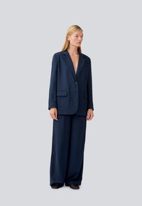 Navy modrý dobře padnoucí blazer a kalhoty s širokými nohavicemi, vyrobené z hladkého materiálu s jedním knoflíkem, hlubokým V-výstřihem a postranními kapsami.