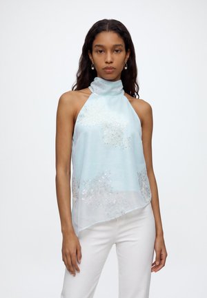 Vrouw met lang donker haar die een lichtblauw haltertopje met zilveren bloemenborduursel en een witte broek draagt, staand tegen een effen achtergrond.