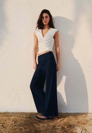 Femme debout contre un mur clair, portant un haut blanc sans manches, un pantalon large bleu marine et des tongs noires sur un sol sec.