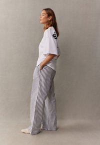 Žena stoji bočno s rukama u džepovima, nosi bijelu široku majicu i široke hlače sivo-bijelih pruga.
