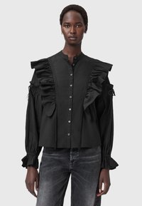 Donna che indossa una camicia nera con bottoni e dettagli arricciati sul petto e sulle maniche, abbinata a jeans neri sbiaditi su uno sfondo chiaro.
