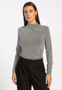 Zilveren langemouwen top met een hoge kraag, gemaakt van een gestructureerde, sprankelende stof. Gecombineerd met hoge zwarte broeken versierd met embellishments.