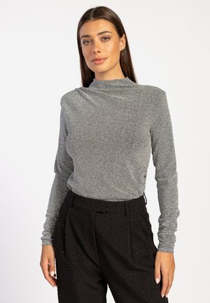 Zilveren langemouwen top met een hoge kraag, gemaakt van een gestructureerde, sprankelende stof. Gecombineerd met hoge zwarte broeken versierd met embellishments.