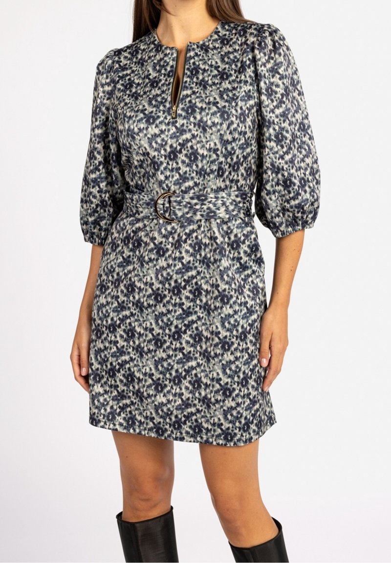 Femme portant une robe ceinturée à motifs bleus et blancs avec des manches trois-quarts bouffantes et un zip à l'avant, associée à des bottes noires hautes jusqu'aux genoux.