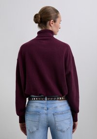 Woman with brown hair in a bun wearing a maroon turtleneck sweater and light blue jeans with a black studded belt, facing away.