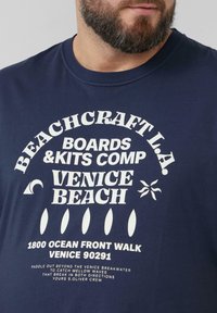 Marineblauwe katoenen t-shirt met gedurfde witte grafische tekst en strandgerelateerde motieven, met een ontspannen pasvorm. Ronde halslijn en korte mouwen.