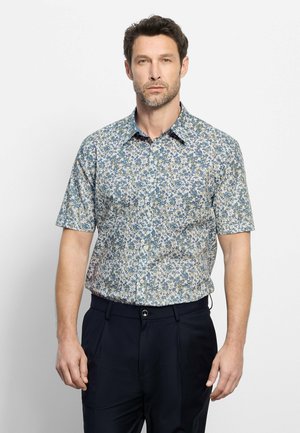 Homme portant une chemise à manches courtes boutonnée avec un motif floral bleu et vert, associée à un pantalon bleu marine foncé.
