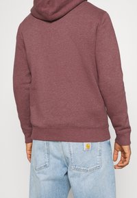 Persona che indossa una felpa con cappuccio rosso scuro e jeans di denim azzurro chiaro con un'etichetta del marchio Carhartt visibile sulla tasca posteriore destra.