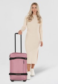 Rosa väska på hjul med svarta remmar och blixtlås, utrustad med ett stadigt handtag och en liten sidoficka, mot en neutral bakgrund.