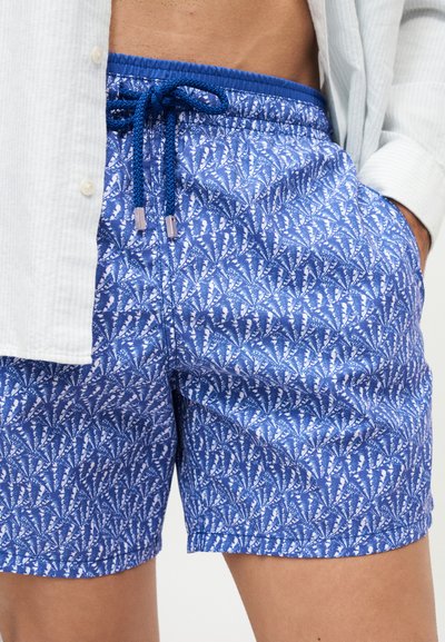Homme portant un short de bain bleu à motifs avec un cordon, une chemise blanche déboutonnée, la main dans la poche et le ventre découvert.