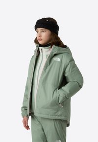 Αδιάβροχο μπουφάν ανοιχτού πράσινου χρώματος με κουκούλα, φερμουάρ σε αντίθετο ροζ χρώμα και λογότυπο. Φοριέται πάνω από λευκό και πράσινο στρώμα ρουχισμού, που συνοδεύεται από υφασμάτινη μάλλινη κορδέλα κεφαλής με υφή.