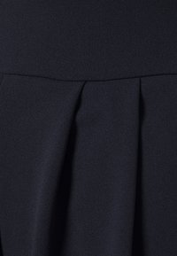 Mörk marinblå klänning i texturerad tyg, med veck vid midjan, slät yta och en figursydd silhuett.