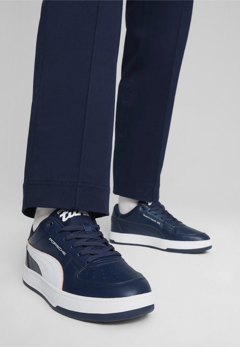 Baskets bleu marine avec semelles blanches et logo Puma blanc portées avec un pantalon bleu foncé et des chaussettes blanches sur fond gris.