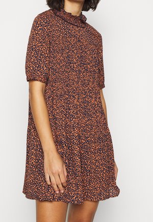 Woman wearing a short-sleeve, above-knee dress with a navy and orange small leaf pattern and a ruffled neckline.