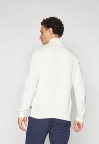 Suéter de cor creme com gola alta, punhos canalé e corte relaxado. Apresenta uma etiqueta de logotipo discreta na bainha e uma textura suave.