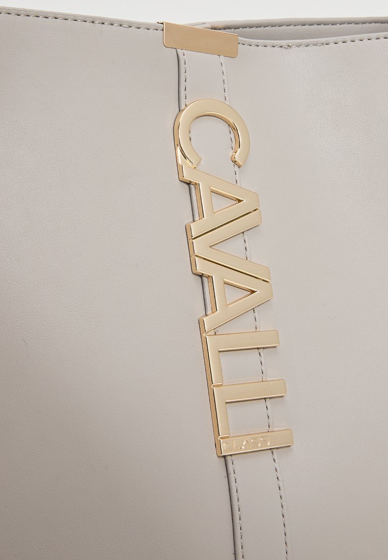 Detail einer beigefarbenen Lederhandtasche mit einem vertikalen, goldfarbenen Metalllogo, das "CAVALLI CLASS" liest und entlang der genähten Naht befestigt ist.