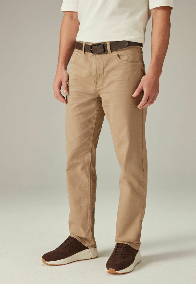 Tan katoenen broek met een slanke pasvorm, voorzien van voorzakken en een bruine riem. Draagt een wit shirt met donkerbruine suède sneakers.