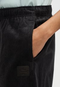 Χέρι που έχει τοποθετηθεί στην πλαϊνή τσέπη μαύρου παντελονιού κοτλέ με ορατές κάθετες ραβδώσεις και μικρή τετράγωνη ετικέτα μάρκας.