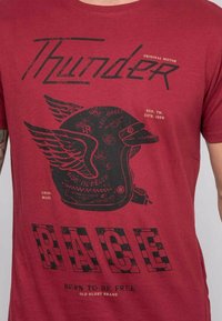T-shirt en coton rouge avec un imprimé graphique noir d'un casque avec des ailes et le texte "THUNDER RACE", incluant une texture altérée et un style vintage.