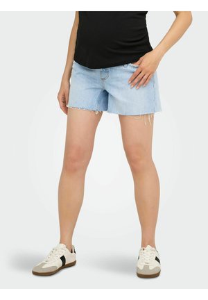 Person iført lyseblå slidte denimshorts, sort top og hvide sneakers med sorte striber, stående mod en hvid baggrund.