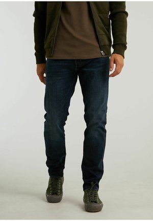 Homme portant un jean bleu foncé, des baskets vert olive, une chemise marron et une veste vert olive ouverte, sur un fond uni.