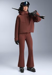 Smučarska obleka v temno rjavi barvi, ki vključuje skrajšan jopič in široke hlače. Vključuje rokavice in smučarske očala. Tekstura iz trpežne tkanine.