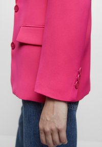 Gros plan sur le bras d'une personne vêtue d'un jean bleu portant un blazer rose vif avec des boutons sur la manche et la patte de la poche avant.