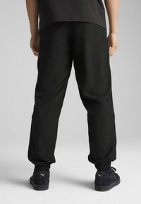 Pantalones negros con cintura elástica y puños fruncidos, y bolsillos laterales, combinados con zapatillas negras que llevan un logo dorado. Tejido suave y ligero.