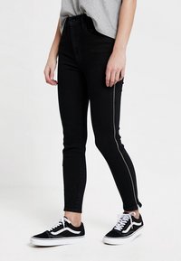 Vaqueros negros ajustados de cintura alta, con detalles de cremallera en las costuras exteriores, combinados con zapatillas negro y blancas. Camiseta gris metida por dentro.