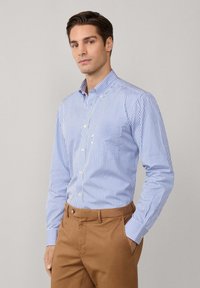 Camisa de manga larga a rayas azules y blancas, con cuello, botones pequeños y un bolsillo en el pecho, combinada con pantalones marrones. Textura de tela suave.