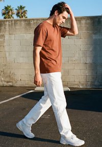 Młody mężczyzna z ciemnymi włosami spacerujący na zewnątrz, ubrany w rdzawy t-shirt, białe spodnie i białe trampki, przeczesujący ręką włosy przy betonowej ścianie.