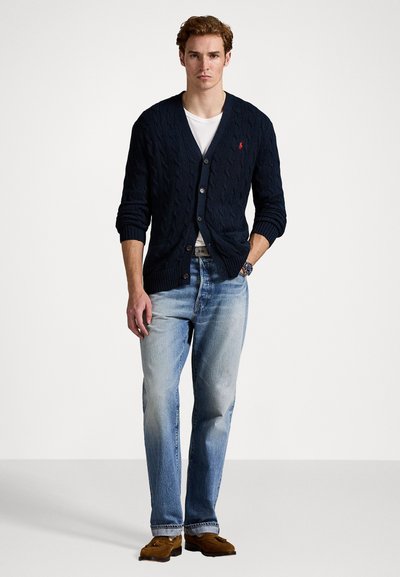 Ναυτικό κασκόλ με πλέξη καλωδίου και σχέδιο V, με κουμπιά, φορεμένο πάνω από άσπρη μπλούζα, συνδυασμένο με ανοιχτό γαλάζιες τζιν και καφέ μοκασίνια.