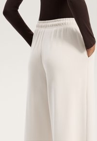 Világos bézs színű, bő szárú nadrág elasztikus derékkal, sima textúrával és oldalsó zsebekkel, egy testhez simuló barna toppal párosítva.