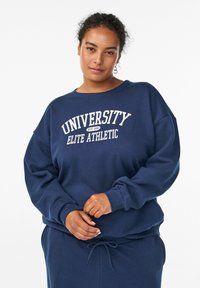 Sudadera azul marino hecha de un tejido suave, con cuello redondo, hombros caídos y texto blanco que dice "UNIVERSITY ELITE ATHLETIC."