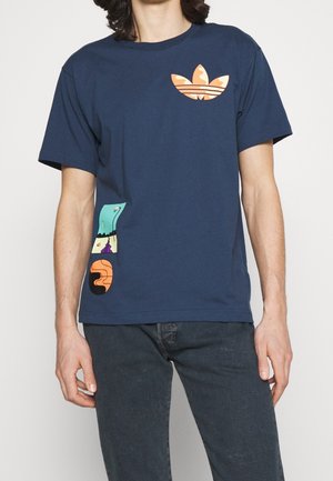 Mężczyzna noszący granatowy t-shirt z pomarańczowym logo Adidas trefoil na piersi i kolorowymi graficznymi kształtami przy lewej talii, w połączeniu z ciemnymi jeansami.