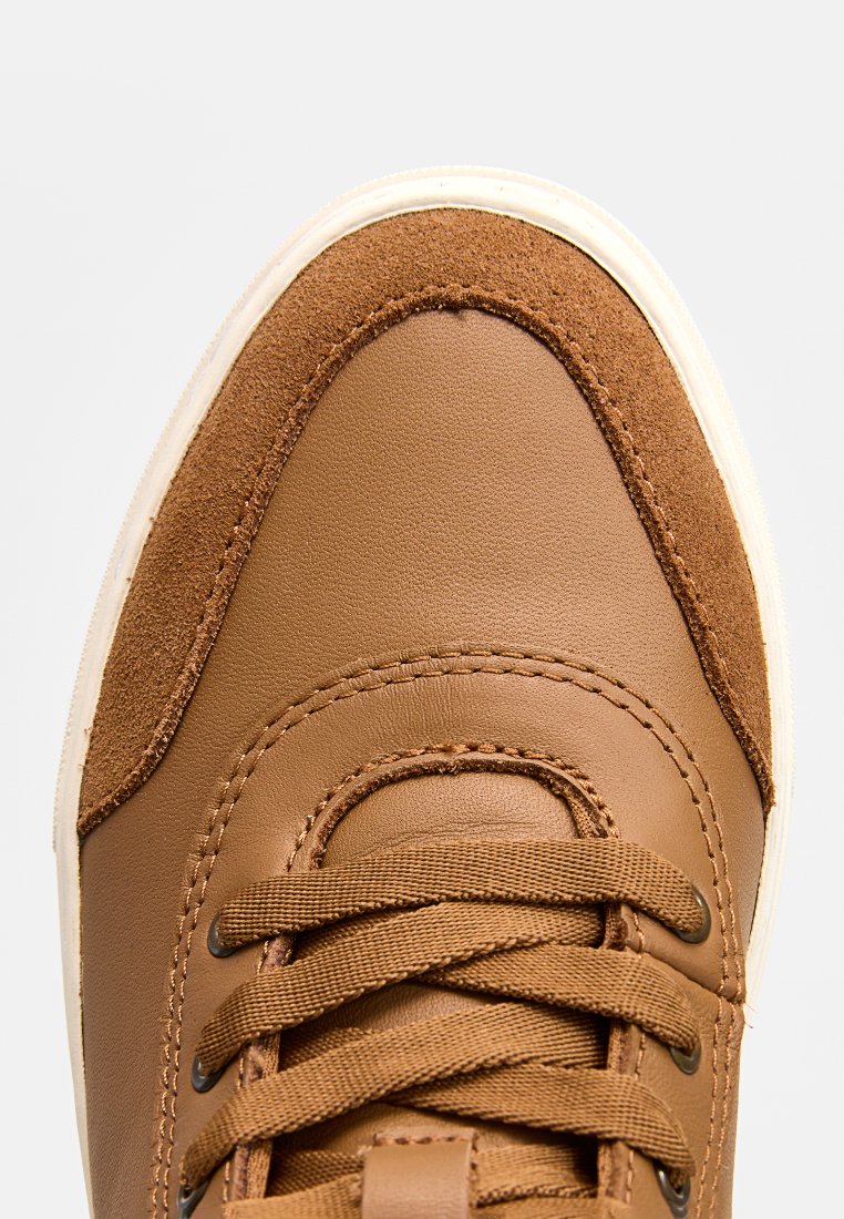 Bruine leren sneaker met een suède neuskap, witte rubberen zool en gewaxt veterdesign. Vloeiende en zachte oppervlaktedetails zijn zichtbaar.