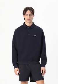 Polo de color azul marino con cuello, que presenta tres botones y un pequeño logo verde en el lado izquierdo del pecho. La tela parece suave y ligeramente texturizada.