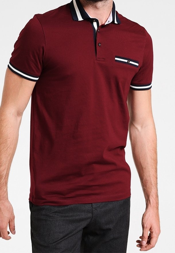 Homme portant un polo bordeaux ajusté avec un col et des poignets de manches rayés noirs et blancs, assorti à un pantalon foncé.