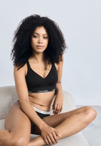 Crni pamukasti bralet s V-izrezom, s elastičnim pojasom s logotipom. Model pozira sjedeći na svijetloj teksturiranoj površini.
