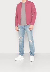 Ljusrosa bomberjacka, grå T-shirt, rivna ljusblå jeans och vita sneakers. Jackan har dragkedja framtill och sidofickor.