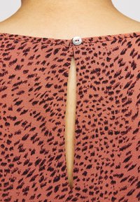 Hnědooranžový top s černými tečkovanými vzory, který má na zádech klíčovou dírku zajištěnou knoflíkem, vyrobený z texturované látky.