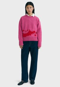 Ung kvinde med langt mørkt hår iført en lyserød og rød sweater over en hvid skjorte, mørkeblå jeans og sorte sko, stående.