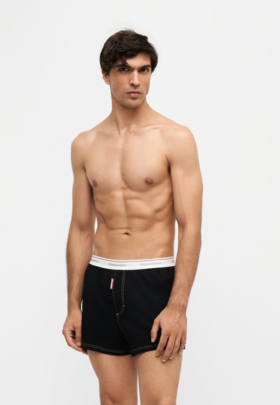 Jeune homme torse nu aux cheveux courts et foncés, portant un boxer noir avec une ceinture blanche, debout devant un fond uni.