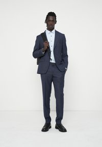 Costum slim-fit de culoare bleu marin, cu un model texturat, cămașă albă și pantofi negri. Subiectul poartă o geantă neagră pe umăr.