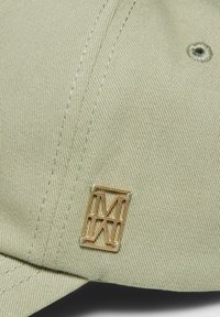 Beige pet met structuurstof, gebogen rand en een gouden metalen logo accent aan de zijkant. Zichtbare stiksels voegen detail toe.